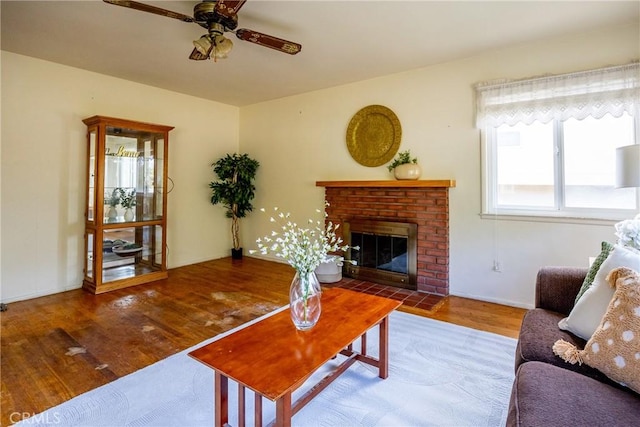 living area featuring a ceiling fan, a brick fireplace, baseboards, and wood finished floors