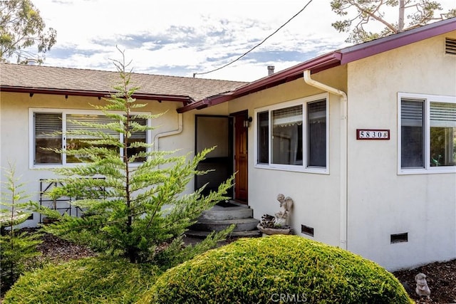 entrance to property featuring a shingled roof, crawl space, and stucco siding