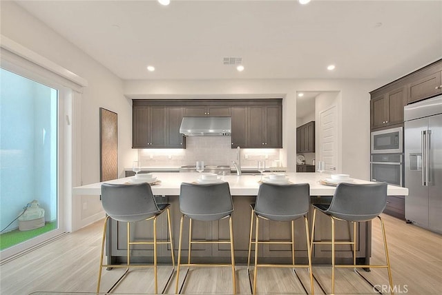 114 Source, Irvine, CA 92618 - kitchen featuring built in appliances, under cabinet range hood, tasteful backsplash, dark brown cabinets, and light wood-style flooring