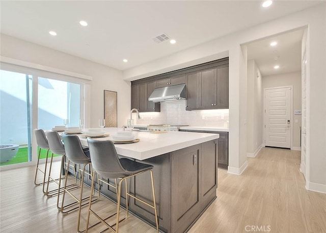114 Source, Irvine, CA 92618 - kitchen featuring under cabinet range hood, decorative backsplash, an island with sink, a breakfast bar area, and light wood-style floors