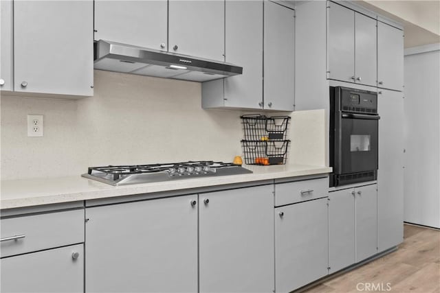 kitchen with light wood finished floors, stainless steel gas cooktop, light countertops, under cabinet range hood, and black oven