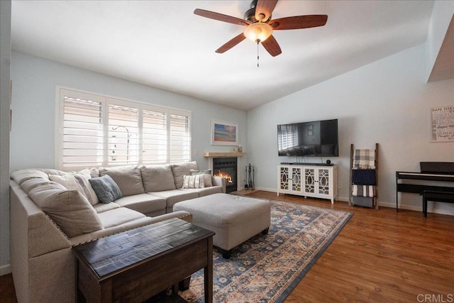 living room with a warm lit fireplace, baseboards, ceiling fan, wood finished floors, and vaulted ceiling