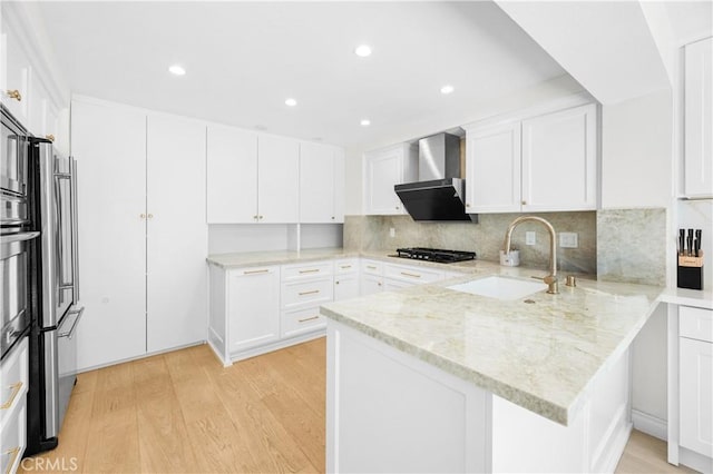 kitchen featuring white cabinets, a peninsula, light wood-type flooring, wall chimney range hood, and a sink