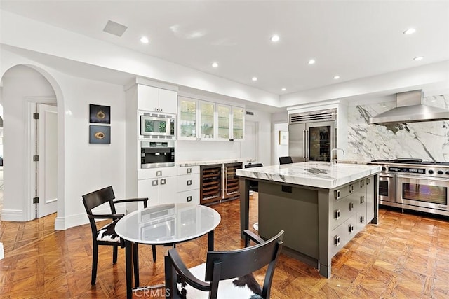 kitchen with built in appliances, arched walkways, white cabinets, wall chimney range hood, and backsplash