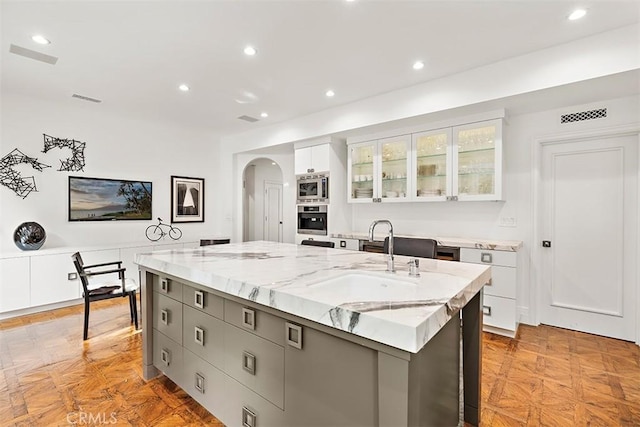 kitchen featuring arched walkways, a kitchen island with sink, stainless steel appliances, white cabinetry, and a sink