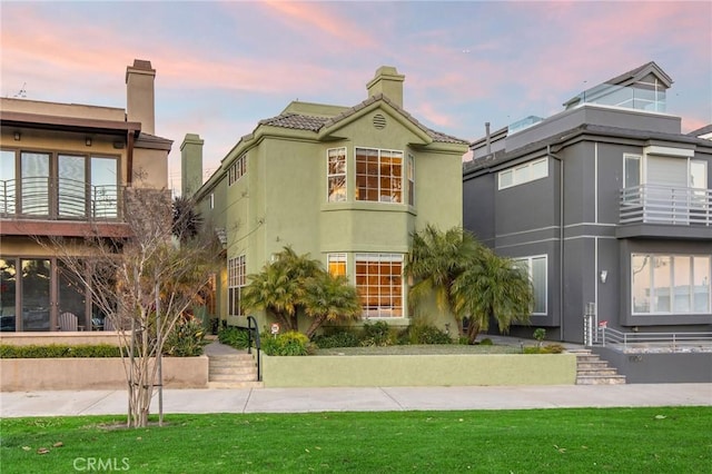 view of front of house with a balcony, a tile roof, stucco siding, a front lawn, and a chimney