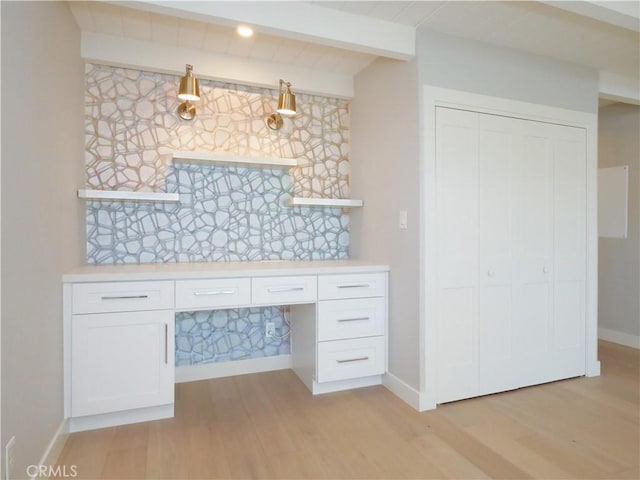 unfurnished office featuring baseboards, built in desk, beamed ceiling, and light wood-style floors