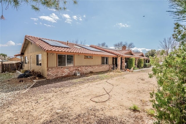 back of house featuring brick siding, fence, a tiled roof, roof mounted solar panels, and a chimney