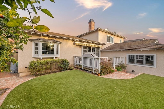 back of property at dusk with stucco siding, a wooden deck, a chimney, and a yard