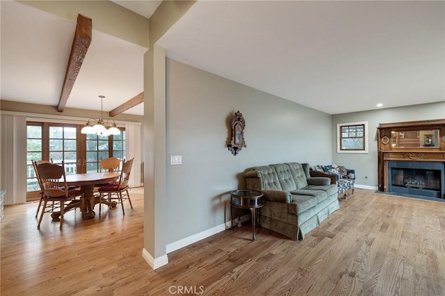 living room with a notable chandelier, light wood-style flooring, a glass covered fireplace, beamed ceiling, and baseboards