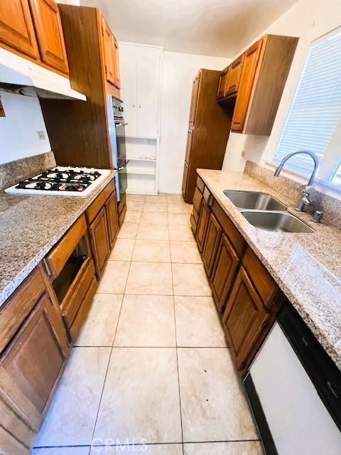 kitchen with light tile patterned floors, a sink, under cabinet range hood, dobule oven black, and white gas stovetop