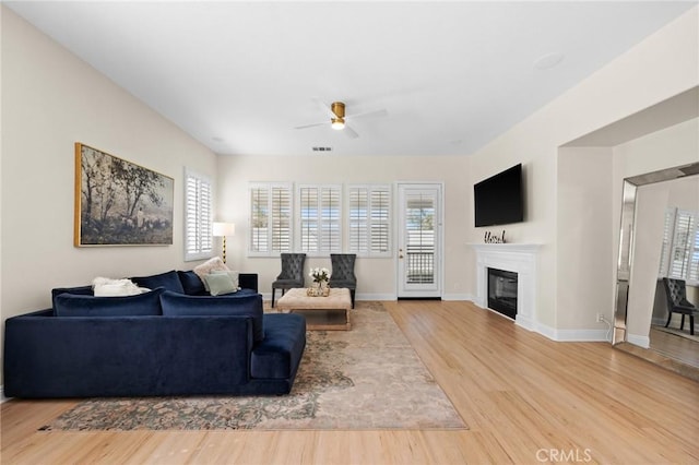 living area with light wood finished floors, visible vents, a glass covered fireplace, ceiling fan, and baseboards