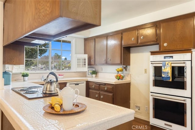 kitchen with brown cabinets, tile countertops, a peninsula, white appliances, and under cabinet range hood