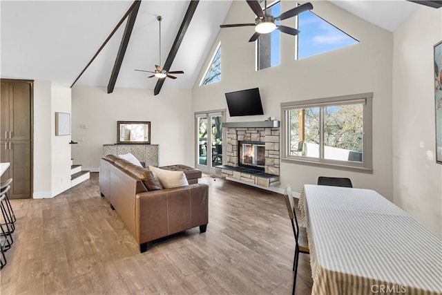 living room with light wood-style flooring, beamed ceiling, a stone fireplace, and a ceiling fan