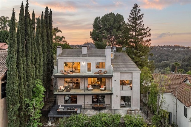 back of house at dusk with a patio, a chimney, a balcony, and stucco siding