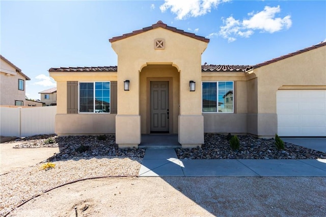 view of exterior entry with an attached garage, a tile roof, fence, and stucco siding