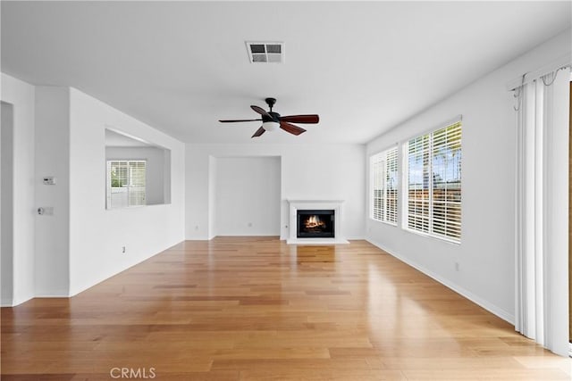 unfurnished living room featuring a warm lit fireplace, ceiling fan, visible vents, baseboards, and light wood finished floors