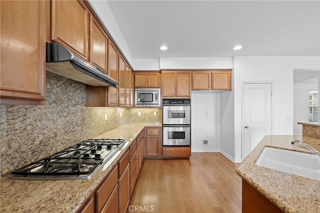 kitchen with decorative backsplash, brown cabinetry, stainless steel appliances, under cabinet range hood, and a sink