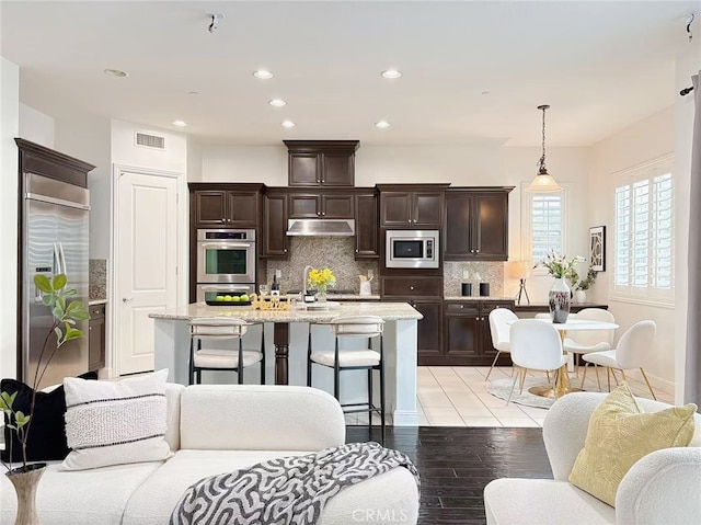 kitchen featuring built in appliances, dark brown cabinetry, under cabinet range hood, visible vents, and backsplash