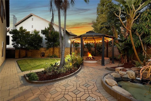 patio terrace at dusk with a fenced backyard, a fire pit, and a gazebo