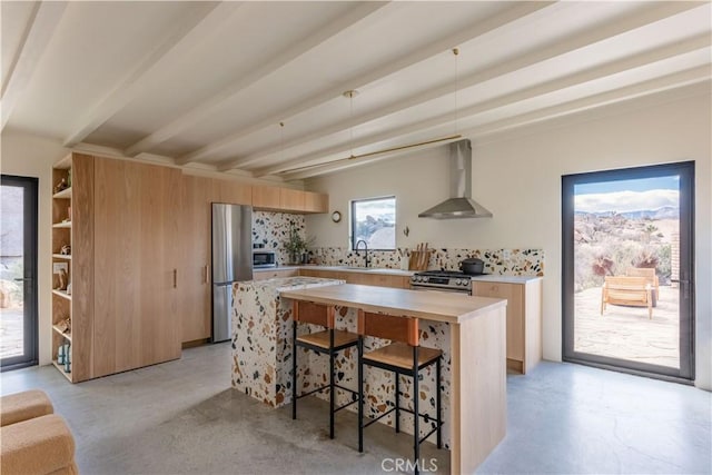 kitchen with beam ceiling, stainless steel appliances, light countertops, wall chimney range hood, and a sink