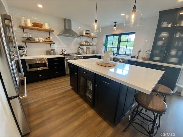 kitchen featuring wall chimney range hood, dark cabinets, appliances with stainless steel finishes, wood finished floors, and open shelves