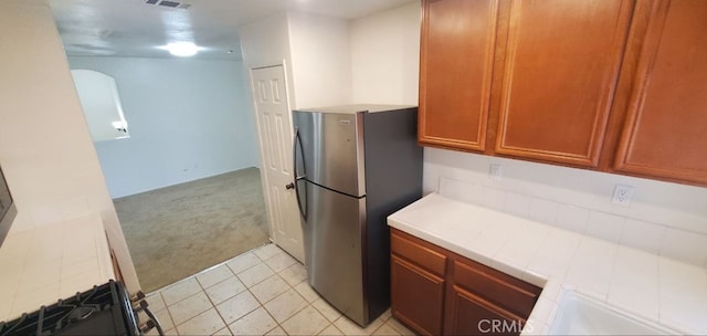 kitchen with tile countertops, light colored carpet, light tile patterned floors, brown cabinets, and freestanding refrigerator