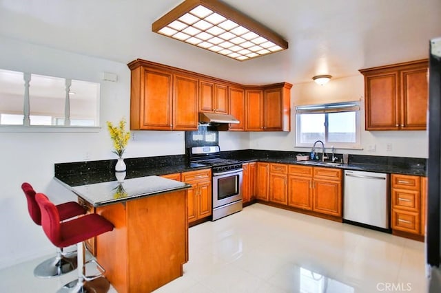 kitchen featuring under cabinet range hood, stainless steel appliances, a peninsula, a sink, and brown cabinets