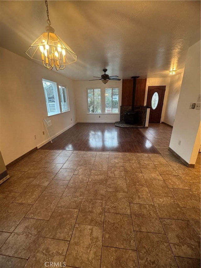 unfurnished living room with ceiling fan with notable chandelier, a wood stove, and baseboards