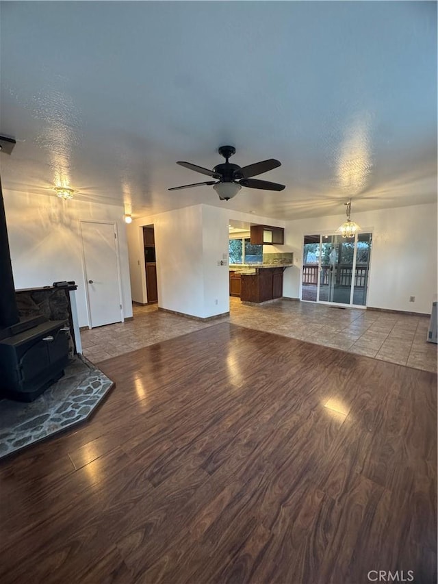 unfurnished living room with ceiling fan with notable chandelier, a wood stove, baseboards, and wood finished floors