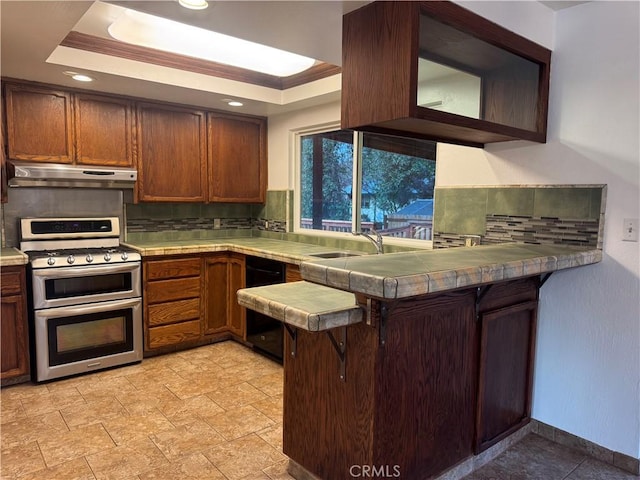 kitchen with a tray ceiling, tile counters, a sink, double oven range, and under cabinet range hood