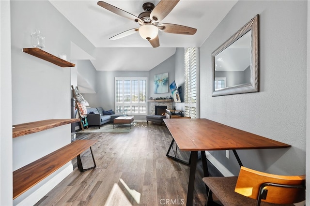 home office featuring a warm lit fireplace, ceiling fan, lofted ceiling, wood finished floors, and baseboards