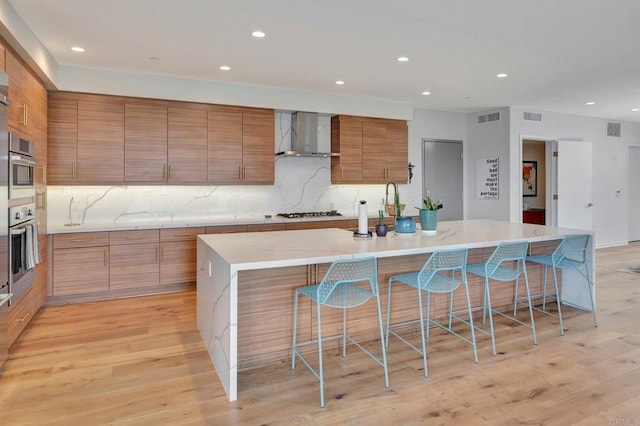 kitchen with brown cabinets, wall chimney exhaust hood, visible vents, modern cabinets, and a kitchen bar