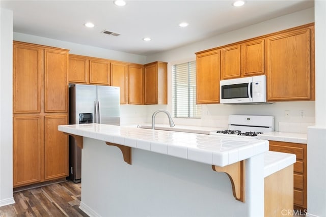 kitchen featuring a sink, a center island with sink, tile countertops, white appliances, and dark wood-style flooring