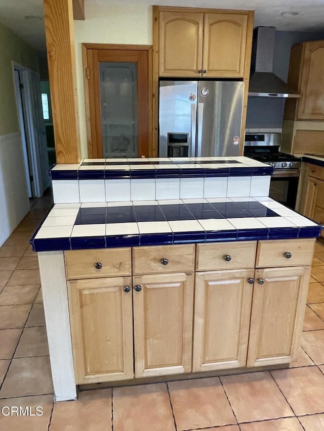 kitchen featuring tile countertops, light brown cabinetry, appliances with stainless steel finishes, and wall chimney range hood