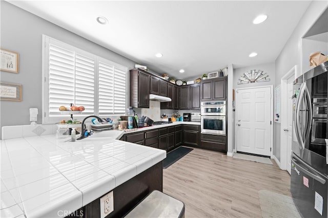 kitchen with appliances with stainless steel finishes, dark brown cabinets, light wood-style floors, a sink, and recessed lighting