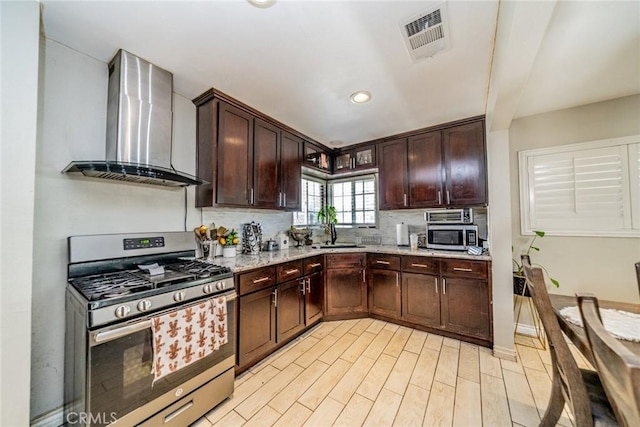 kitchen featuring dark brown cabinetry, stainless steel appliances, visible vents, wall chimney exhaust hood, and tasteful backsplash