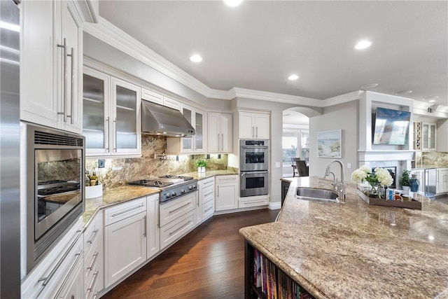 kitchen with under cabinet range hood, appliances with stainless steel finishes, arched walkways, white cabinetry, and a sink