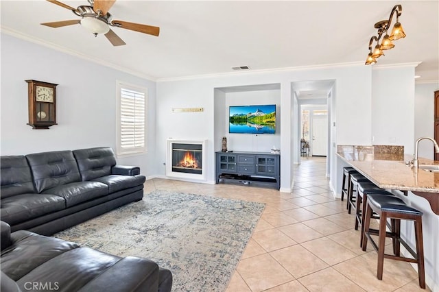 living room with light tile patterned floors, visible vents, a ceiling fan, a glass covered fireplace, and crown molding