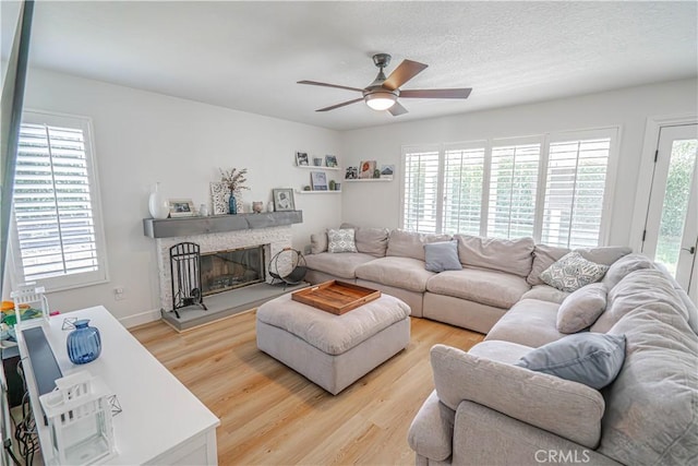 living area with light wood finished floors, a ceiling fan, a stone fireplace, a textured ceiling, and baseboards