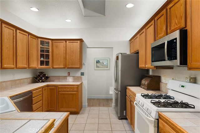 kitchen with tile countertops, stainless steel appliances, recessed lighting, brown cabinetry, and glass insert cabinets