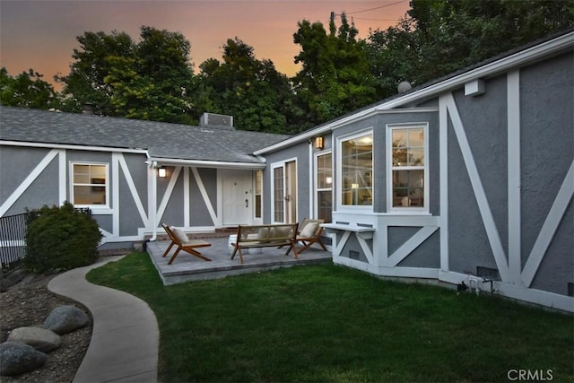 back of house at dusk with a yard, a shingled roof, a patio area, and stucco siding