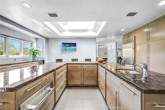 kitchen featuring a skylight, built in refrigerator, a peninsula, a tray ceiling, and a sink