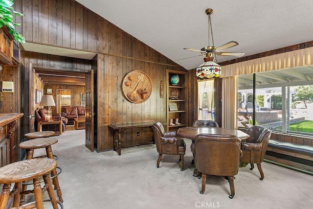 dining room with ceiling fan, wooden walls, vaulted ceiling, and light colored carpet