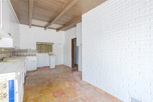 interior space with beam ceiling, tile countertops, decorative backsplash, white cabinets, and brick wall