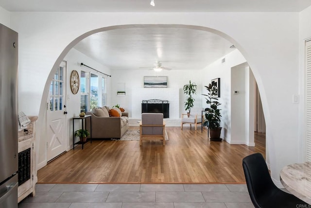 living room featuring light tile patterned floors, ceiling fan, a brick fireplace, and arched walkways