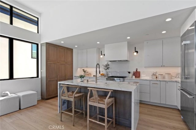 kitchen featuring premium range hood, high end refrigerator, a sink, light wood-style floors, and a center island with sink