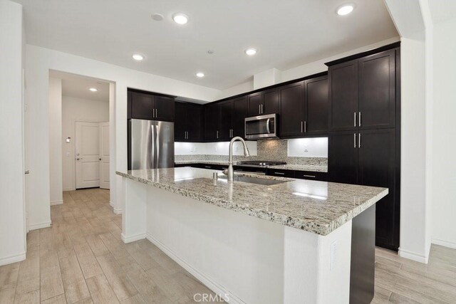 kitchen featuring a kitchen island with sink, stainless steel appliances, a sink, light stone countertops, and light wood finished floors