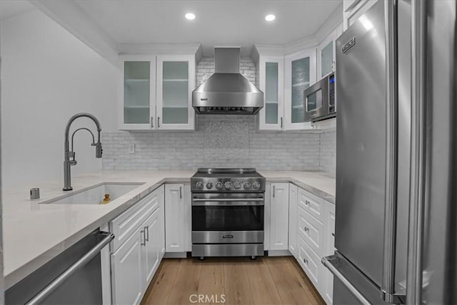 kitchen with stainless steel appliances, light countertops, a sink, wall chimney range hood, and wood finished floors