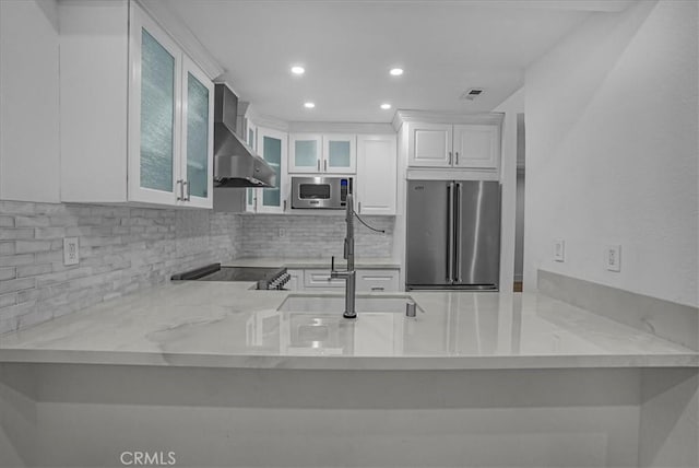kitchen featuring a peninsula, backsplash, wall chimney exhaust hood, stainless steel appliances, and a sink